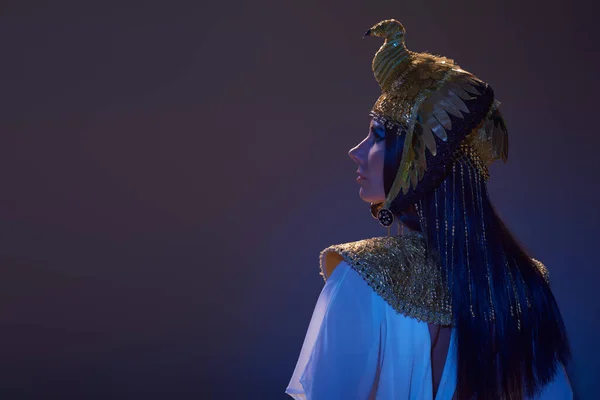 Vista lateral de la mujer con estilo en tocado egipcio de pie sobre fondo marrón con luz azul - foto de stock