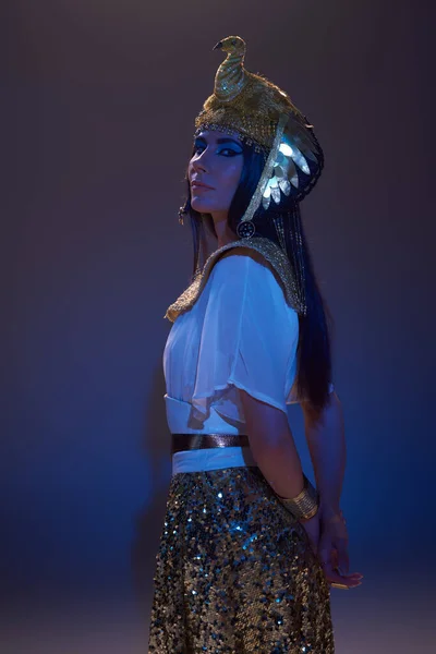 Mujer elegante en aspecto egipcio dorado y tocado mirando a la cámara en marrón con luz azul - foto de stock