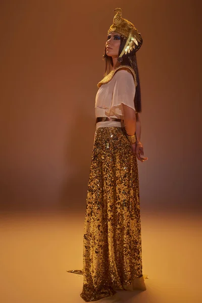 Повна довжина жінки в золотому єгипетському погляді і головний убір, що стоїть на коричневому фоні — стокове фото