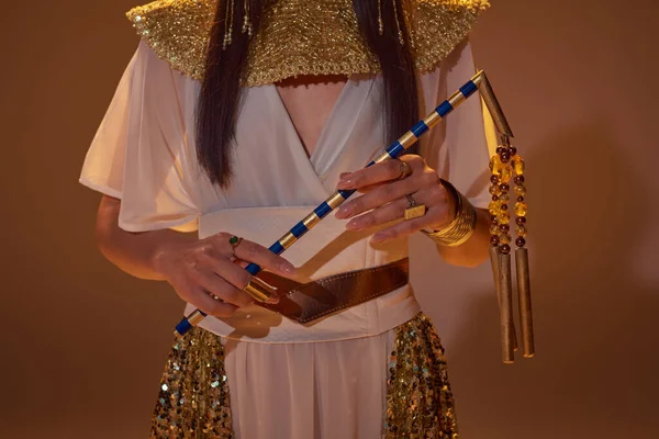 Ausgeschnittene Ansicht einer Frau im ägyptischen Outfit, die Dreschflegel hält, während sie auf braunem Hintergrund posiert — Stockfoto