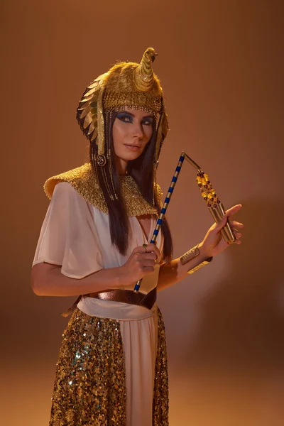 Schöne Frau in ägyptischer Kleidung und Kopfschmuck hält Dreschflegel, während sie auf braunem Hintergrund posiert — Stockfoto