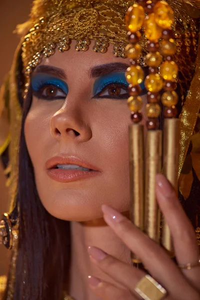 Porträt einer eleganten Frau mit kühnem Make-up und ägyptischer Kopfbedeckung, die Dreschflegel isoliert auf braun hält — Stockfoto