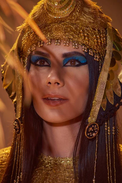 Porträt einer Frau mit elegantem ägyptischen Kopfschmuck, die in der Nähe von Wüstenpflanzen steht, isoliert auf braun — Stockfoto