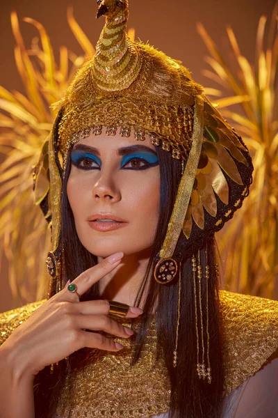 Mujer elegante en aspecto egipcio y tocado posando cerca de plantas borrosas sobre fondo marrón - foto de stock