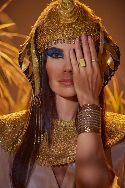 Жінка в елегантному єгипетському вбранні, що закриває обличчя і стоїть біля пустельних рослин ізольовано на коричневому — стокове фото