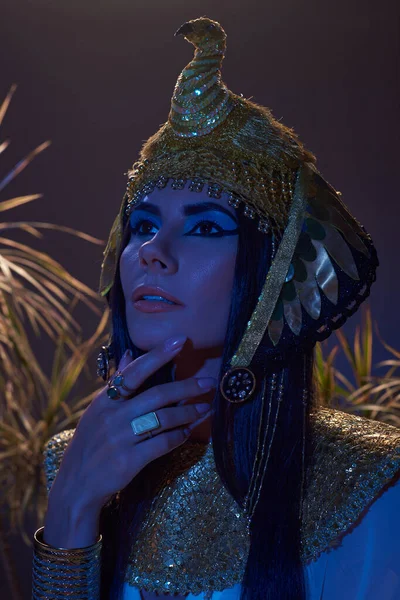 Porträt einer Frau mit ägyptischem Kopfschmuck, die in der Nähe von Pflanzen in blauem Licht auf braunem Hintergrund wegschaut — Stockfoto