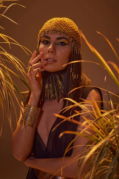 Porträt einer brünetten Frau mit ägyptischem Blick, die in die Kamera schaut, in der Nähe von Pflanzen auf braunem Hintergrund — Stockfoto
