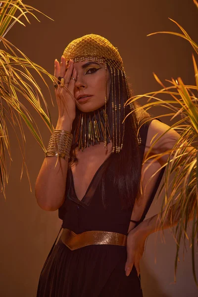 Frau in ägyptischem Outfit und Kopfbedeckung bedeckt Gesicht, während sie neben Pflanzen auf braunem Hintergrund steht — Stockfoto