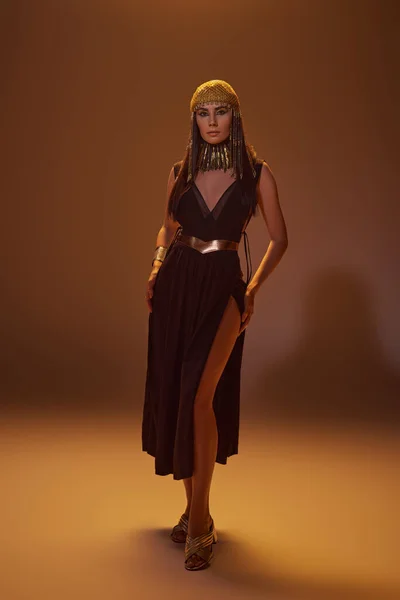 Повна довжина елегантної жінки в єгипетському вигляді і головний убір стоїть і позує на коричневому фоні — стокове фото