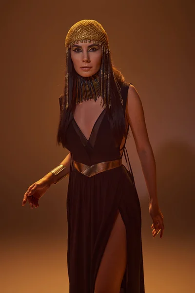 Жінка в єгипетському вигляді і намисто дивиться на камеру, стоячи на коричневому фоні зі світлом — стокове фото