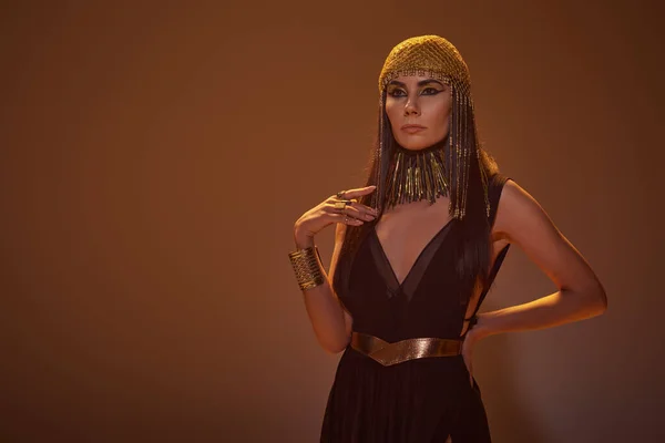 Femme à la mode en look égyptien et collier tenant la main sur la hanche tout en se tenant debout sur fond brun — Photo de stock