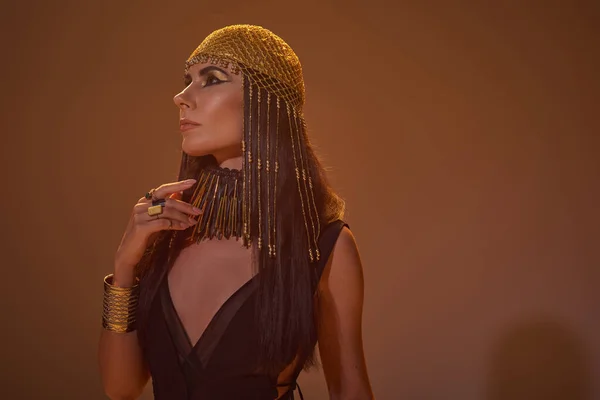 Mujer elegante con maquillaje audaz y traje egipcio de pie sobre fondo marrón con luz - foto de stock