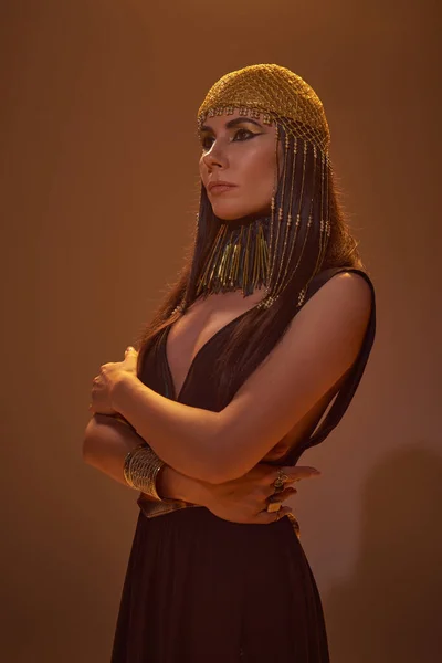 Mujer de moda en mirada egipcia y tocado posando y mirando hacia otro lado sobre fondo marrón - foto de stock