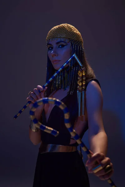 Stylish woman in egyptian attire holding flail and crook in blue light on brown background — Stock Photo