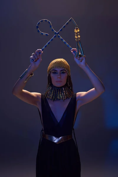 Mulher com maquiagem egípcia e olhar segurando vigarista e flail no fundo marrom com luz azul — Fotografia de Stock