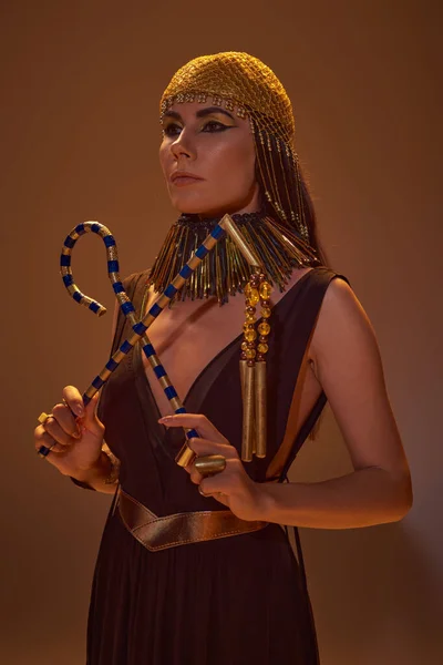 Elegante mujer en tocado dorado egipcio y mirada sosteniendo ladrón y flail sobre fondo marrón - foto de stock