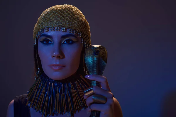 Frau mit ägyptischer Kopfbedeckung und Make-up hält Gauner in Schlangenform auf blauem Licht auf braun — Stockfoto