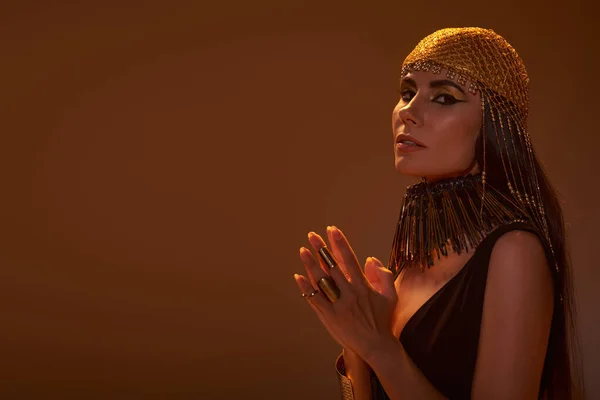 Femme en coiffe dorée et regard égyptien regardant la caméra isolée sur brun avec de la lumière — Photo de stock