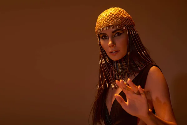 Mulher em cobertura para a cabeça egípcia e olhar posando para a câmera no fundo marrom com luz — Fotografia de Stock