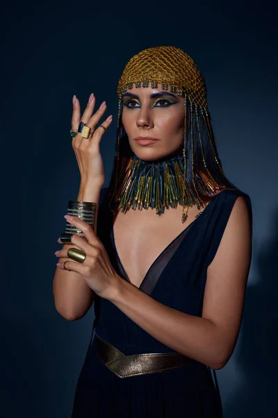 Mujer elegante en mirada egipcia y collar posando mientras está de pie sobre fondo azul - foto de stock