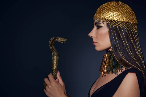 Side view of woman in egyptian attire holding crook in snake shape on blue background — Stock Photo