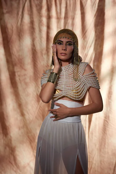 Modelo elegante no olhar egípcio e pérola topo tocando bochecha e de pé sobre fundo abstrato — Fotografia de Stock