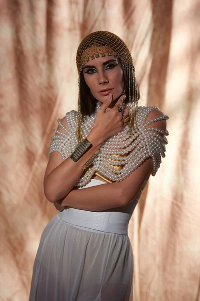Mujer morena de moda en mirada egipcia y top de perlas mirando a la cámara sobre fondo abstracto - foto de stock