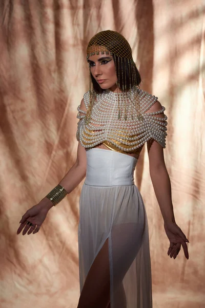 Mujer atractiva con maquillaje audaz posando en traje egipcio y top de perlas sobre fondo abstracto - foto de stock