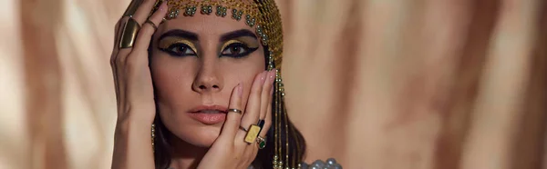 Brünette Frau mit ägyptischem Make-up und Kopfbedeckung, die Gesicht auf abstraktem Hintergrund berührt, Banner — Stockfoto