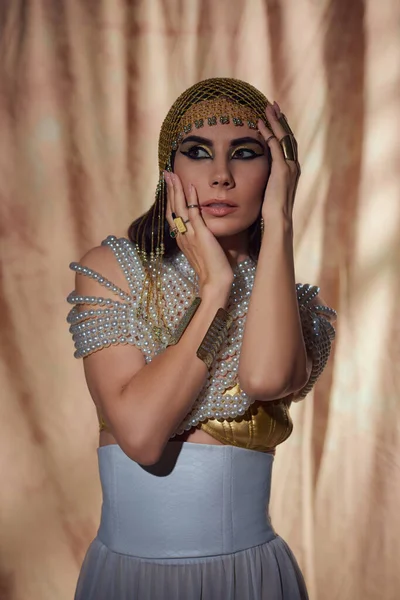 Mulher com maquiagem ousada, cobertura para a cabeça egípcia e top pérola tocando o rosto em fundo abstrato — Fotografia de Stock