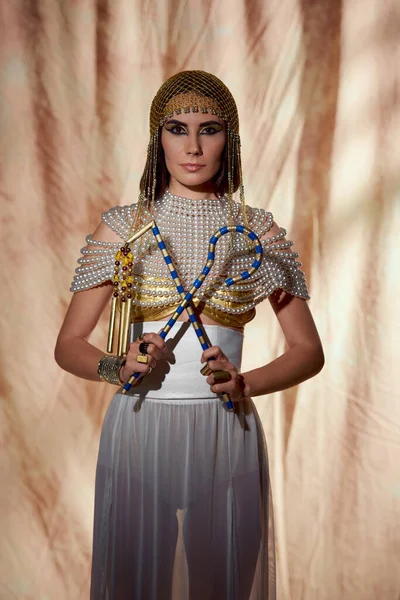 Frau in ägyptischem Kostüm und Perlenkleid hält Gauner und Dreschflegel in der Hand, während sie auf abstraktem Hintergrund posiert — Stockfoto