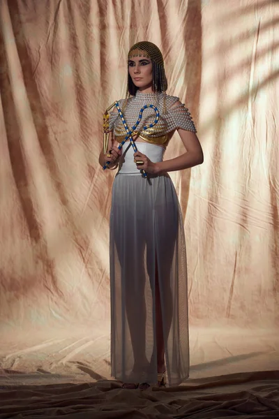 Modische Frau mit ägyptischem Kopfschmuck und Perlenoberteil, die Dreschflegel und Gabel auf abstraktem Hintergrund hält — Stockfoto