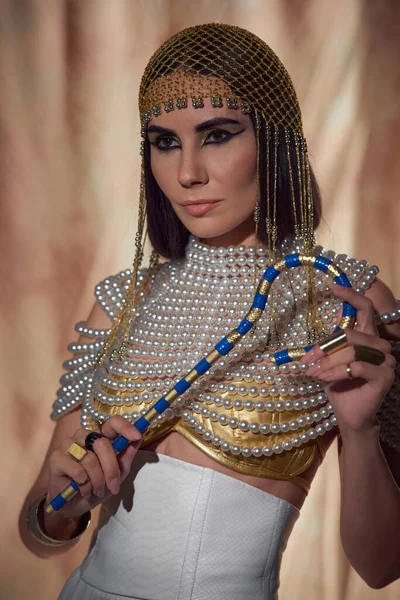 Элегантная женщина в головном уборе и перламутровом топе держит египетского мошенника на абстрактном фоне — стоковое фото