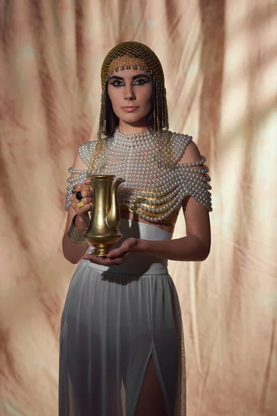 Elegant woman in egyptian look and pearl necklace holding shiny jug on abstract background — Stock Photo