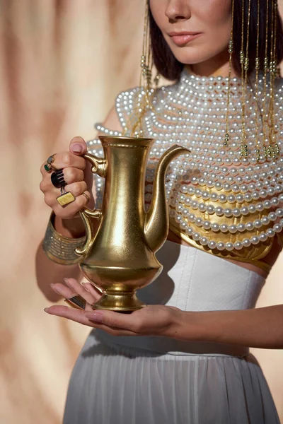 Обрізаний вид стильної жінки в єгипетському погляді, що тримає золотий глечик на абстрактному тлі — стокове фото
