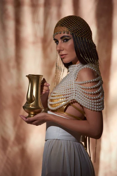 Элегантная женщина в египетском головном уборе и перламутровом топе держит золотой кувшин на абстрактном фоне — стоковое фото
