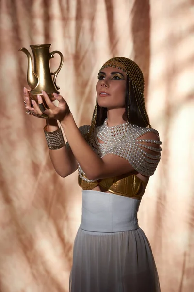 Femme élégante en tenue égyptienne et haut perlé regardant la cruche dorée sur fond abstrait — Photo de stock