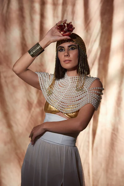 Стильна жінка в єгипетському вигляді і сміливий макіяж, що тримає вирізаний гранат на абстрактному фоні — стокове фото