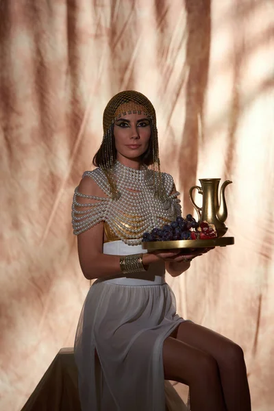 Mulher elegante em trajes egípcios e top pérola segurando jarro e frutas frescas em fundo abstrato — Fotografia de Stock