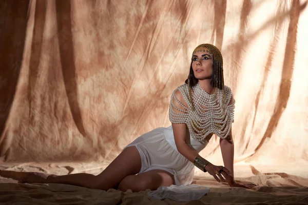 Brünette Frau in ägyptischem Look und Perlentop, die auf abstraktem Hintergrund mit Sonnenlicht wegschaut — Stockfoto