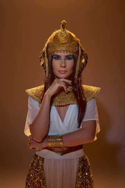Stilvolle brünette Frau mit Make-up und ägyptischer Kleidung schaut in die Kamera auf braunem Hintergrund — Stockfoto