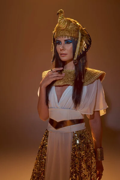 Brünette Frau in ägyptischer Kopfbedeckung und stilvollem Look posiert auf braunem Hintergrund mit Beleuchtung — Stockfoto
