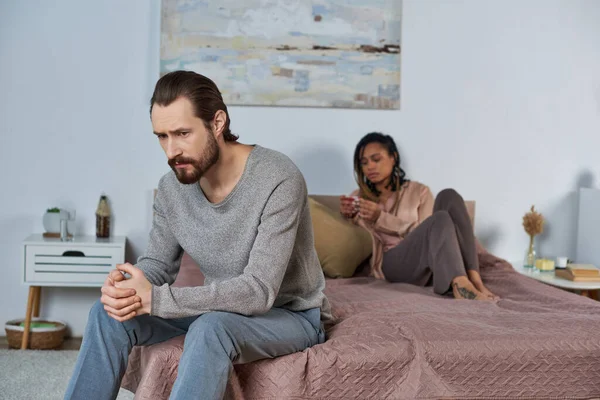Homme inquiet assis sur le lit, se sentant stressé, femme afro-américaine avec test de grossesse, décision — Photo de stock