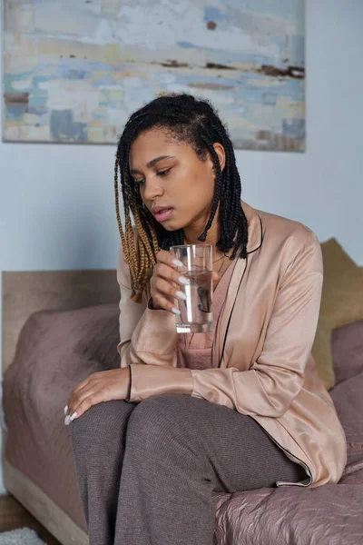 Zugeschnittene, traurige Afroamerikanerin sitzt auf dem Bett, fühlt sich unwohl, hält ein Glas Wasser in der Hand, Schlafzimmer — Stockfoto