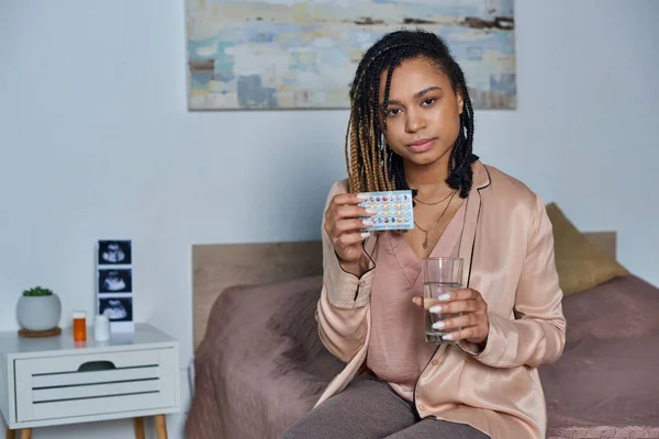 Афроамериканська жінка тримає склянку води і таблетки контролю народжуваності, вагітність, сучасна спальня — стокове фото