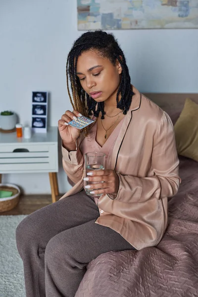 Афроамериканська жінка тримає склянку води і таблетки контролю народжуваності, вагітність, сидячи в спальні — стокове фото