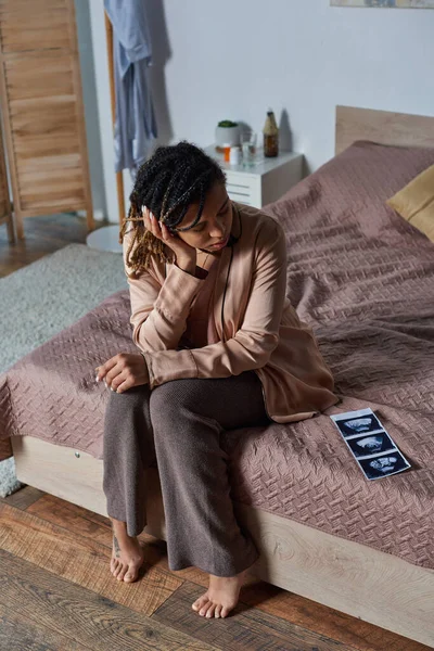 Mujer afroamericana sentada en la cama y mirando la foto de ultrasonido, vaso de agua, control de natalidad - foto de stock