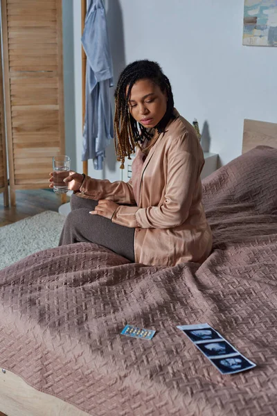 Ultrasonido y píldoras anticonceptivas en la cama, mujer afroamericana sentada con un vaso de agua, cama - foto de stock