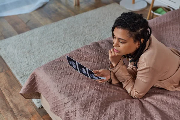 Top view of worried african american woman lying on bed, looking at ultrasound, unborn, abortion — Stock Photo
