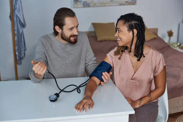 Happy bearded man measuring blood pressure of pregnant african american wife, future parents, tattoo — Stock Photo